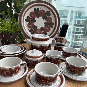 Könnte beinhalten: Ein Keramik-Teeservice mit weißem Untergrund und einem braun-dunkelorangen Frucht- und Blattmuster. Das Set besteht aus einer Teekanne mit Deckel, einer Zuckerdose mit Deckel, einem Milchkännchen, mehreren Tassen mit Untertassen und einer großen Servierplatte.