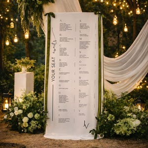 Puede incluir: Un plano de asientos de tela blanca con detalles de cinta verde y el texto "YOUR SEAT awaits". El gráfico enumera los nombres alfabéticamente, perfecto para una boda o evento. El fondo presenta vegetación e iluminación suave.