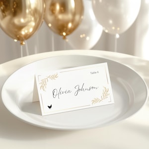 May include: A white place card with gold accents, featuring the name "Olivia Johnson" in elegant script and a small black chicken illustration. The card is set on a white plate, with gold and white balloons in the background.