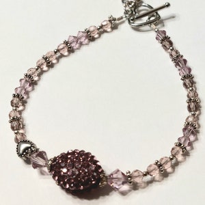 May include: A delicate bracelet with a heart-shaped clasp and a large, sparkly pink bead in the center. The bracelet is made with small, clear and pink glass beads.