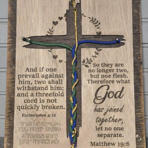 May include: Wooden cross-shaped wall art with a braided cord design in green, blue, and gold. The cross is set in a rustic wooden frame. The artwork features Bible verses and decorative elements.