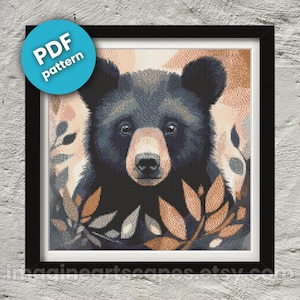 May include: A black bear cub with brown eyes peeks out from behind a leafy branch. The bear's fur is dark and glossy, and its nose is pink. The background is a soft, blurred orange and brown.