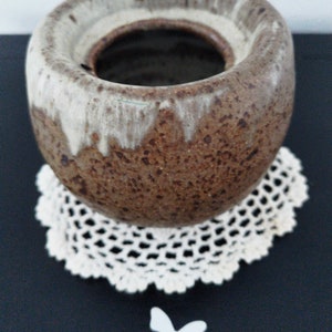 May include: A brown and white ceramic vase with a rough texture. The vase has a wide opening and a rounded shape. It is sitting on a white crocheted doily.