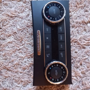 Puede incluir: Un panel de control de clima de coche negro con un borde plateado y botones. El panel tiene una pantalla digital, una perilla de control de temperatura y un botón de control de zona. El texto "Avantgarde" es visible en el panel.