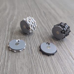 Puede incluir: Cuatro placas de peso magnéticas grises y blancas y dos ajustadores de peso cilíndricos se muestran sobre una superficie de madera. Las placas tienen el número 5. Los ajustadores son grises y blancos o grises y negros.