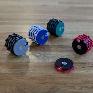 Peut inclure: Quatre compteurs cylindriques en forme d'engrenage avec des aimants centraux, composés de pièces en plastique noir et coloré. Un compteur est gris, un bleu, un turquoise et un rouge. Un compteur circulaire plat rouge et noir est au premier plan.