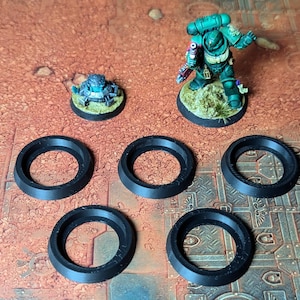 Puede incluir: Una vista aérea muestra figuras en miniatura y bases de plástico negro. Una figura verde y turquesa se encuentra sobre una base circular con hierba. Se ven cinco bases circulares negras con un borde biselado. También hay una figura más pequeña.