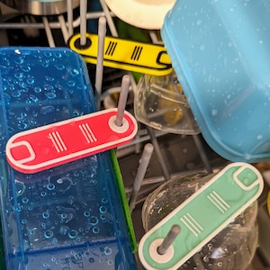 May include: Close-up of a dishwasher rack with three colourful plastic bottle holders. The holders are red, yellow, and green and have three white lines on each. The holders are attached to metal rods.