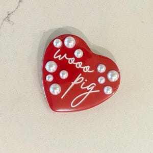 May include: A red heart-shaped object with the words "wooo pig" in white script. The heart is adorned with several white pearl-like embellishments. The object is likely a decorative item or accessory.