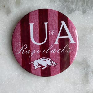 May include: A round button with a maroon and white striped background. The button features the text "UA of Razorbacks" in white, along with a white illustration of a Razorback hog.