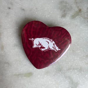 May include: A heart-shaped trinket box with a maroon finish and a white Razorback logo. The box has a glossy surface and a smooth, rounded shape. The logo is a stylized image of a running wild boar.