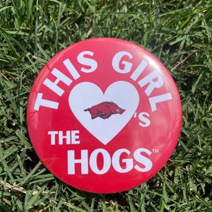 May include: A red button with white text that reads "THIS GIRL'S THE HOGS" in a circular pattern. A white heart in the center contains a Razorback logo. The button is set against a background of green grass.