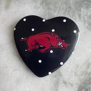 May include: Heart-shaped button with a black background and white polka dots. A red Razorback hog graphic is in the center. The button is a collectible item.