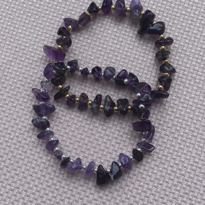 May include: Two amethyst chip bracelets with gold and silver beads. The bracelets are on a pink textured background.