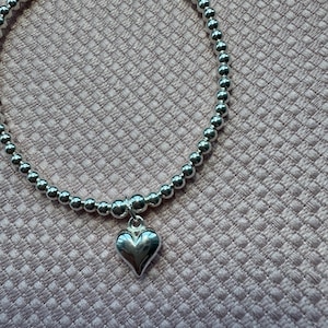 May include: A silver bracelet featuring small, round beads and a heart pendant. The beads are strung together to form a circular shape. The heart charm is smooth and hangs from the centre. The background is a textured, pink fabric.