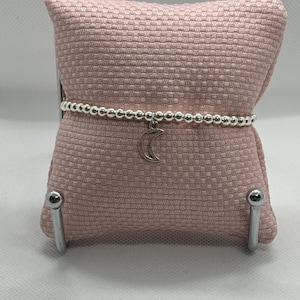 May include: A silver beaded bracelet with a crescent moon charm. The bracelet is displayed on a pink cushion.