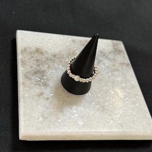 May include: A silver beaded ring with a single white bead on a black ring display stand.