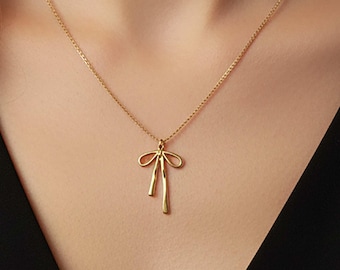 May include: A delicate gold-colored necklace featuring a bow pendant. The bow has a looped design with two dangling vertical bars. The necklace is worn against a black top, highlighting the jewelry's design.