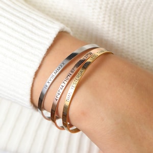May include: Three engraved metal bangles in silver, rose gold, and gold. The silver bangle has the inscription "IX.XVIII.MMXX". The rose gold bangle has the inscription "41°47'44.710"N 87°137"W". The gold bangle has the inscription "MORE THAN YESTERDAY LESS THAN".