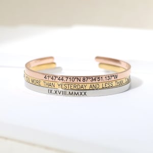 May include: Three stacked gold, rose gold, and silver bangle bracelets with engraved text. The top bracelet reads "41°47'44.710"N 87°34'51.137"W". The middle bracelet reads "You more than yesterday and less than tomorrow". The bottom bracelet reads "IX.XVIII.MMXX".