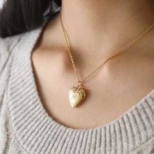 May include: A gold heart-shaped locket necklace with an engraved letter "R" on the front. The necklace is worn by a person with a light brown sweater.