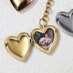 May include: A gold heart-shaped locket with a photo of a woman and a dog inside. The locket is open and the back is visible. The locket is attached to a gold chain.