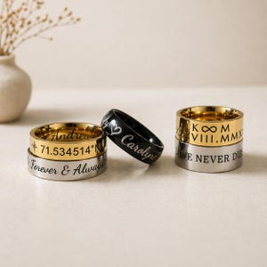 May include: A collection of personalized rings in gold, silver, and black. The rings feature engraved text, including names, coordinates, and phrases like "Forever & Always" and "Love Never Die."