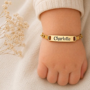 May include: A gold-colored baby bracelet with the name "Charlotte" engraved in black lettering. The bracelet is on a small wrist, with a soft, neutral-colored background. The bracelet has a chain-link design.