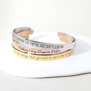 May include: Three stacked bracelets with engraved text. The top bracelet is silver and reads "27°58′19.79″N, 82°30′7.19″W". The middle bracelet is gold and reads "Making Every Moment Matter". The bottom bracelet is rose gold and reads "Be the change that you wish to see in the world".