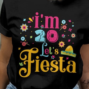 May include: Black t-shirt with a colorful graphic that reads "I'm 20 Let's Fiesta." The design includes the number 20, a sombrero, and floral accents in pink, blue, yellow, and green. The shirt is a casual style.