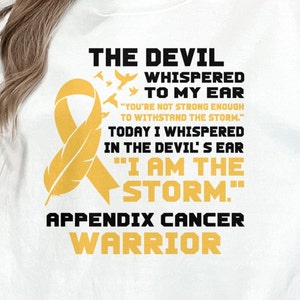 Puede incluir: Camiseta blanca con una cinta dorada y el texto "THE DEVIL WHISPERED TO MY EAR "YOU'RE NOT STRONG ENOUGH TO WITHSTAND THE STORM." TODAY I WHISPERED IN THE DEVIL'S EAR "I AM THE STORM." APPENDIX CANCER WARRIOR"