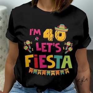 May include: Black t-shirt with the text "I'M 40 LET'S FIESTA" in colorful letters. The design includes a sombrero, maracas, and a banner. The shirt is a crew neck and has short sleeves.