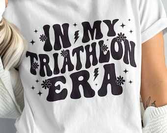 May include: White t-shirt with black text that reads "In My Triathlon Era" in a retro font. The text is surrounded by small black flower and lightning bolt graphics.
