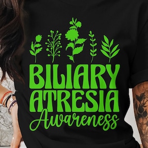 Op de afbeelding: Zwart T-shirt met groene tekst en bloemmotieven. De tekst luidt "Biliary Atresia Awareness" in een retro lettertype. Boven de tekst staan groene bloemillustraties.