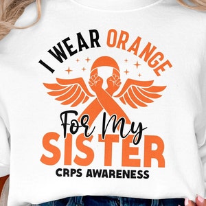 Op de afbeelding: Witte sweatshirt met de tekst "I Wear Orange For My Sister" in zwart en oranje. Het ontwerp bevat een oranje lint met vuisten en vleugels, en de woorden "CRPS Awareness" onderaan.