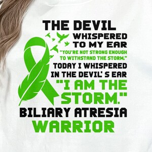 Op de afbeelding: Groen lint met een verenontwerp, een wit T-shirt met de tekst "THE DEVIL WHISPERED TO MY EAR "YOU'RE NOT STRONG ENOUGH TO WITHSTAND THE STORM." TODAY I WHISPERED IN THE DEVIL'S EAR "I AM THE STORM." BILIARY ATRESIA WARRIOR"