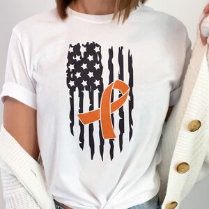 Puede incluir: Camiseta blanca con un diseño de bandera estadounidense desgastada y una cinta naranja en el centro.