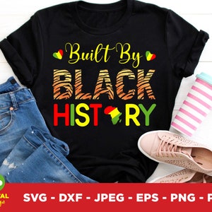 Pode incluir: Camiseta preta com o texto "Built By BLACK HISTORY" em amarelo, laranja, vermelho e verde. O design inclui um mapa da África. A camisa é combinada com jeans azuis, um copo rosa e branco e tênis rosa.