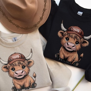 May include: Two sweatshirts with a cartoon highland cow wearing a cowboy hat that says "Howdy". One sweatshirt is beige and the other is black.