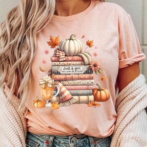 May include: A pink t-shirt with a graphic design of a stack of books, a pumpkin, and a cup of coffee. The text on the shirt reads "Just a girl who loves fall & books".