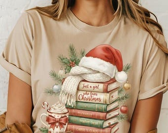May include: Beige t-shirt featuring a Christmas-themed graphic. The design includes a stack of books with titles like "Just a girl who loves Christmas," a Santa hat, a scarf, and a cup of hot cocoa with a candy cane.