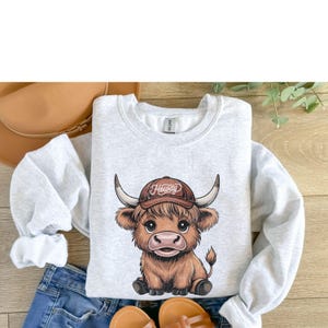 May include: A gray sweatshirt with a cartoon illustration of a brown Highland cow wearing a brown baseball cap with the word "Howdy" on it.
