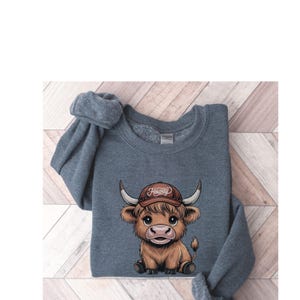May include: A gray sweatshirt with a cartoon illustration of a brown Highland cow wearing a brown baseball cap with the word "Howdy" on it.