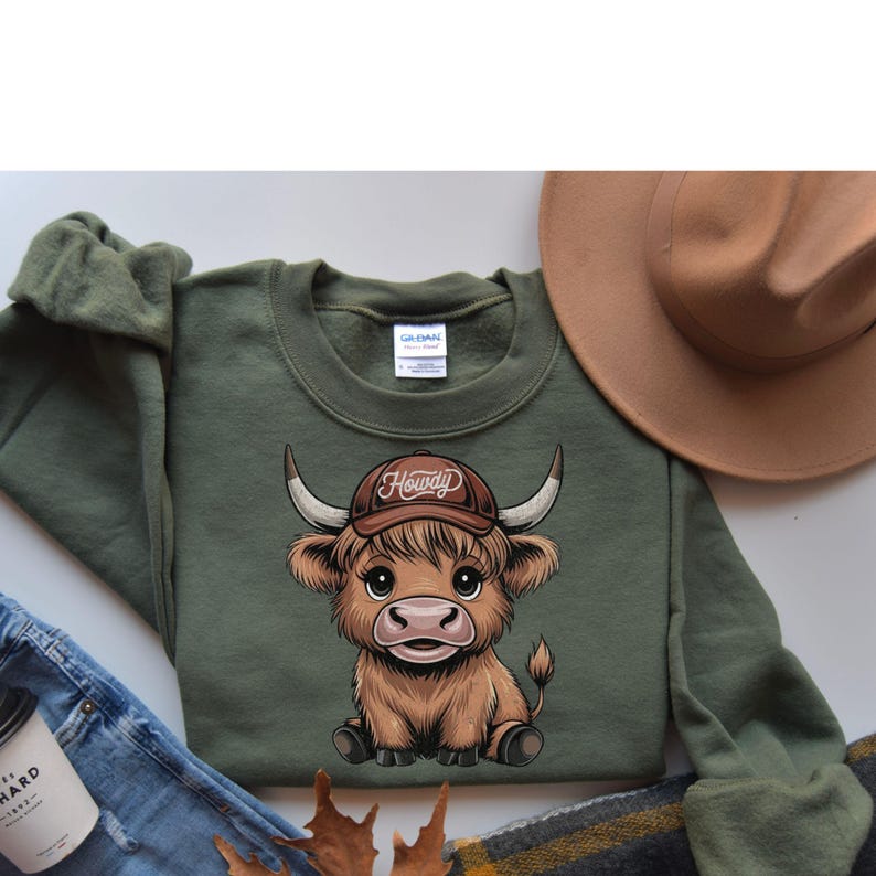 May include: Green sweatshirt with a cartoon illustration of a brown Highland cow wearing a brown cowboy hat with the word "Howdy" on it.