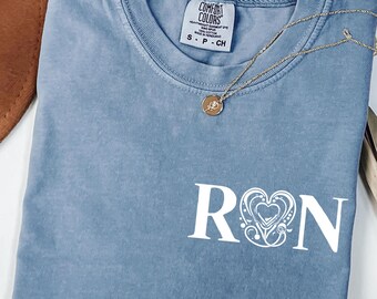 May include: A light blue t-shirt with a white graphic of the letters "RN" with a stylized heart design in between. The shirt has a distressed look and is made of a soft, comfortable fabric. The text "COMFORT COLORS" is visible on the tag.