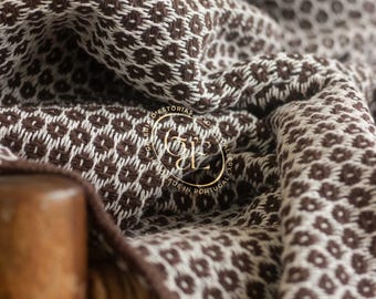 May include: A close-up of a brown and white patterned textile. The fabric is draped over a wooden chair. A gold emblem with text is visible on the fabric. The textile has a repeating geometric design.