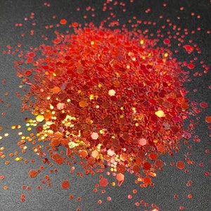 May include: A pile of red glitter with various shapes and sizes, including hexagons and small dots. The glitter has a shiny, reflective surface and is scattered on a dark background. The glitter appears to be a craft supply.