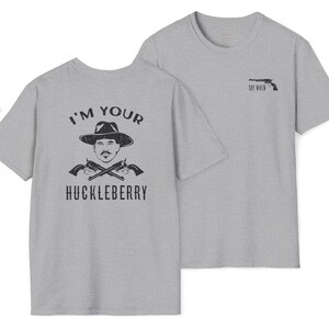 Puede incluir: Dos camisetas grises. Una presenta un gráfico de un hombre con sombrero y pistolas cruzadas con el texto "I'M YOUR HUCKLEBERRY". La otra camiseta tiene un gráfico de pistola y el texto "SAY WHEN."