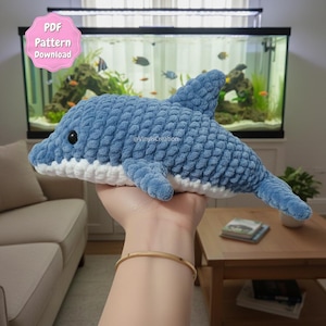 Könnte beinhalten: Ein blaues Plüsch-Delphin-Spielzeug mit weißen Akzenten, das in einer Hand gehalten wird. Der Delphin hat ein strukturiertes, schuppenartiges Muster und ein schwarzes Auge. Das Bild enthält den Text "PDF Pattern Download".