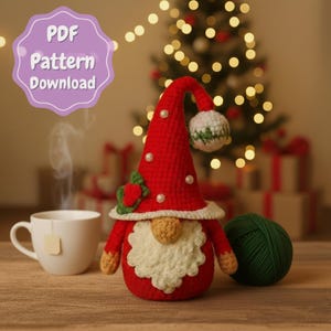 Puede incluir: Un gnomo de ganchillo hecho a mano con un gorro rojo adornado con perlas y una barba blanca. El gnomo tiene un pompón de gorro verde y blanco y un acento floral verde y rojo. Una taza blanca y una madeja de hilo verde están junto al gnomo. La imagen incluye el texto "PDF Pattern Download".